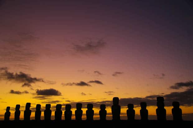 Silhouetted Moai statues with a sunset in the background with clouds and orange skies.