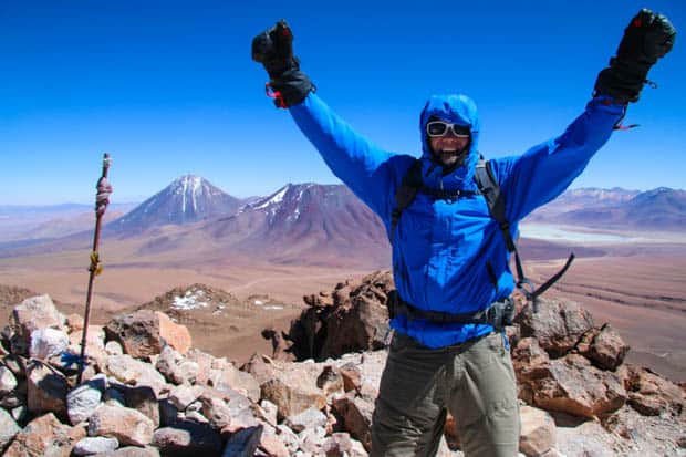 Author on the top of a mountain with arms in the air in celebration of summitting with mountains and plains in the background.