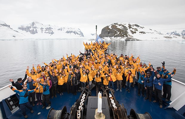 Entire crew and guests posing with their hands up in the air on the bow of their small ship in Antarctica.