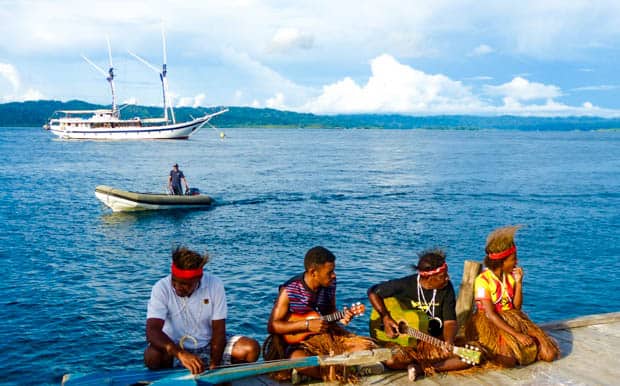Locals playing music on the dock in Indonesia with a small ship in the background.