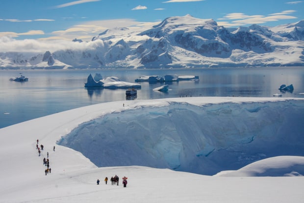 People snowshoeing uphill on Antarctica with their small cruise ship in the background.