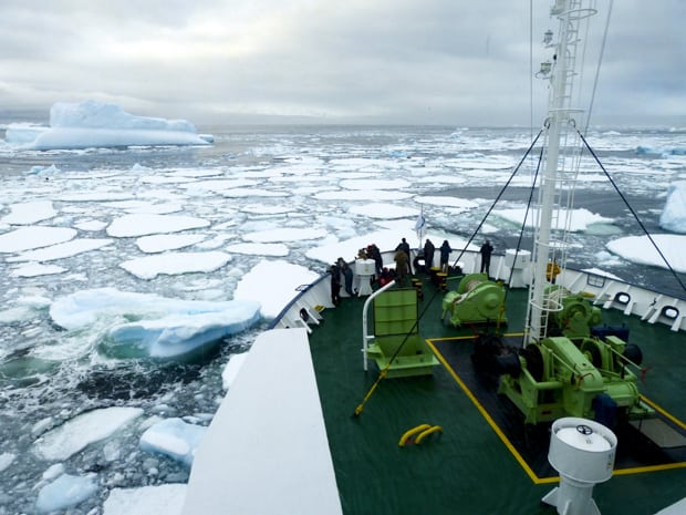 People on the bow of their small ship watching as the ship breaks through some ice in Antarctica.