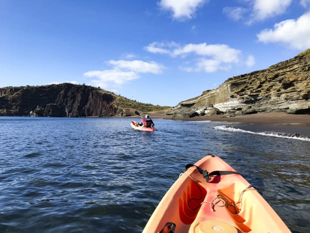 2 kayakers gliding off the shore of a sandy beach with bluffs.