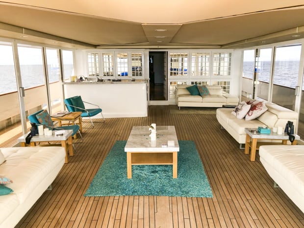 Deck view of a enclosed outdoor lounge area with couches and chairs and a bar. Large windows and sliding glass doors access the deck.