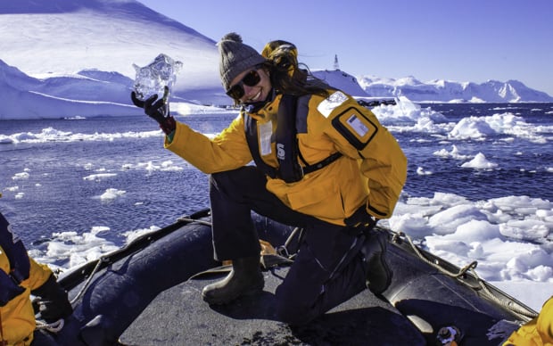 Happy guest from a small ship aboard a skiff excursion posing with a piece of glacial ice with icebergs behind in Antarctica.