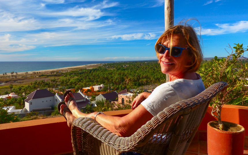 Woman in blue sunglasses sits in white wicker chair overlooking the ocean on a sunny day in Baja California.