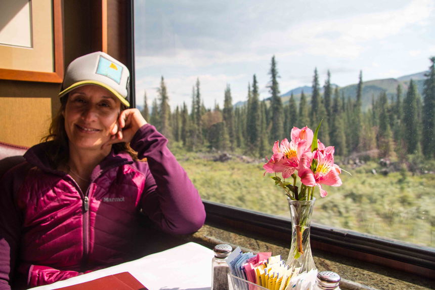 Woman in berry-colored coat sits with head propped on hand at table with pink flowers u0026 view window on Alaska Railroad dining car.