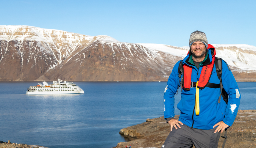 Adventuresmith Andrew posing for a picture with a blue jacket on and a red life vest in front of his aurora small ship cruise on a sunny day in front of snowy mountains
