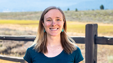 A woman in a dark blue shirt and blue earrings smiles at the camera with a fence behind her