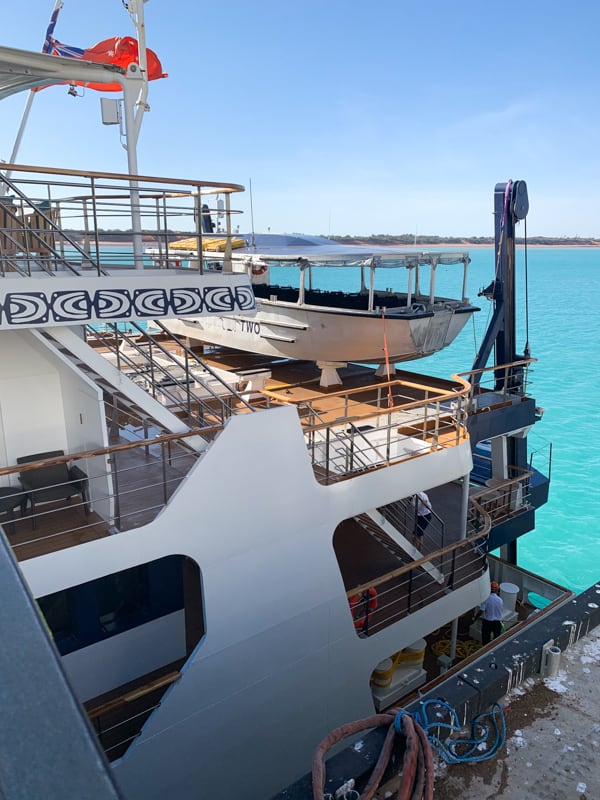 The stern or back of the Coral Adventurer showing three decks and the storage of the small vessel tender, behind is a bright teal ocean and a cloudless sky