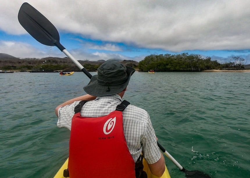 Blue sky and teal water as a kayaker paddling through the ocean in the Galapagos wearing a red life jacket and a grey wide brim ha