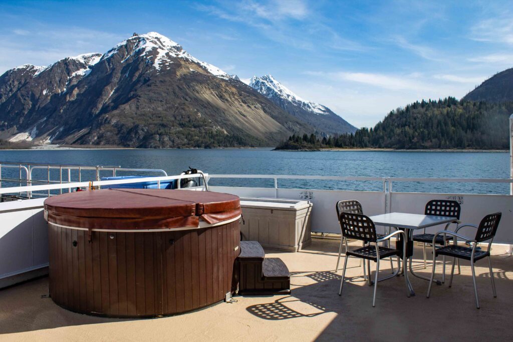 the top deck of small ship Wilderness Adventurer an outdoor table and chairs are set beside a covered Jacuzzi, in the distance a jaged snow capped mountain range end where the water line begins