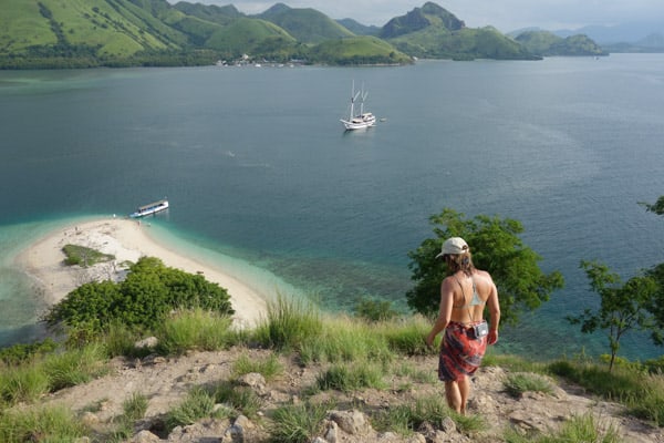 a female traveler walks down a hill side towards a sandy beach, the ocean and views of islands can be seen.