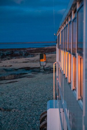 The sun is setting in the Canadian arctic, the orange and pink colors reflect off the windows of the polar rover buss off road vehicle as it is parked in front of the shoreline on the classic polar bear adventure tour 