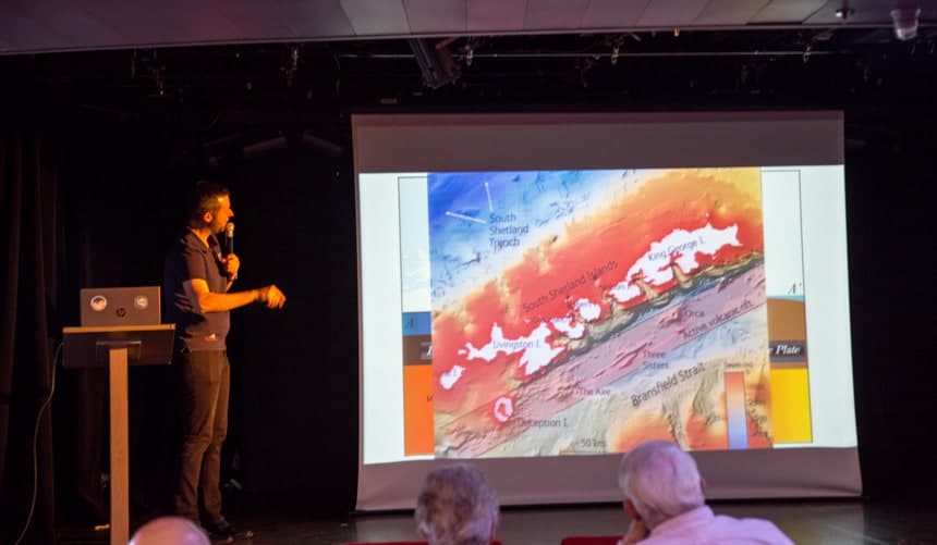 Aboard a luxury small ship, a male speaker stands in front of an audience at a podium holding a laptop computer, narrating an educational video on a screen.