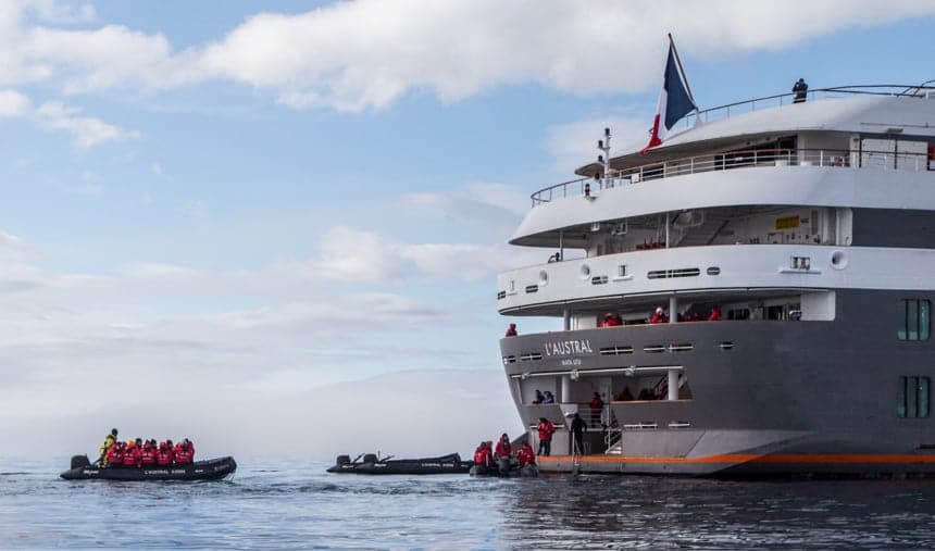 At the stern or back of the luxury ship L'Austral, black inflatable skiffs boat up to a landing dock where passengers in red parkas disembark and get back aboard the ship.