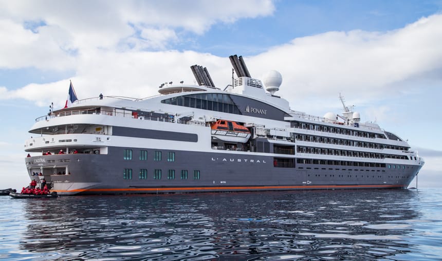 Floating on the blue ocean on a blue cloudy day, luxury ship L'Austral has a blue hull with red stripe, with 4 stories of white decks with private balconies.