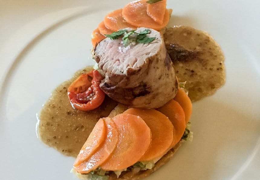 A main course diner plate of meat surrounded by orange carrots, red baby tomatoes with a sauce. Served aboard L'Austral a luxury small ship.