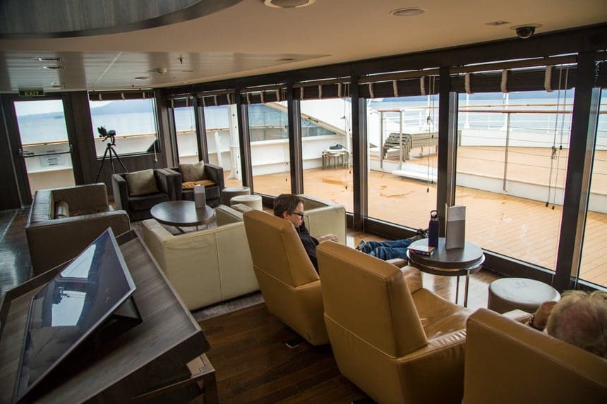 A lounge seating area aboard L'Austral and her ponant luxury sister ships. surrounded with large glass windows cream, beige and brown leather arm chairs are set facing the windows with ottomans and coffee tables.