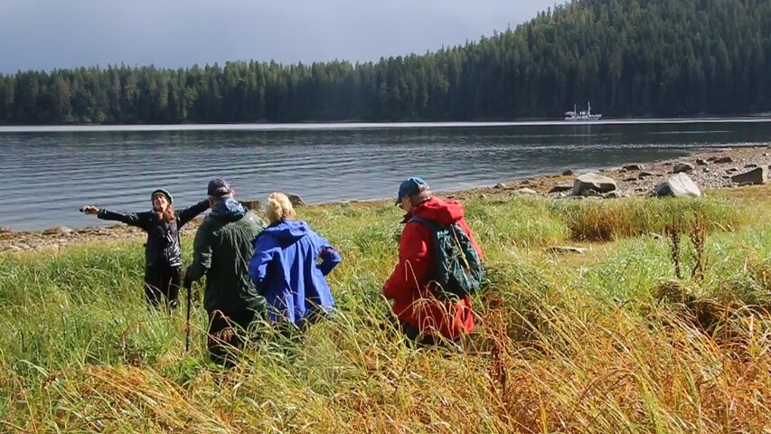 A shore excursion on a small ship Alaska cruise, the naturalist guide opens her arms wide as three guests walk toward her through a grassy field, in the distance small ship catalyst floats on water intron of a forested hillside.