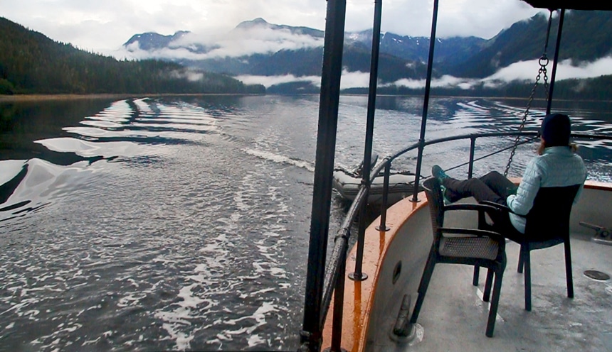 A female traveler sits in a chair on the stern of Catalyst, feet up, drinking coffee looking out at the Alaska landscape.
