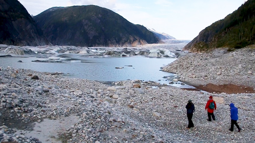3 travelers on a Catalyst cruise shore excursion walk a rocky shoreline surrounded by mountains near a glacier in Alaska