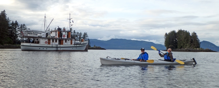 two travelers paddle a gray double kayak in front of the small ship Catalyst on their slow travel cruise in Alaska