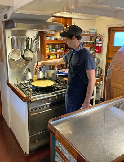 An open kitchen aboard Alaska small ship catalyst, the chef wears a blue apron and holds a spatula in a pan filled with food.