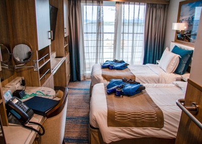Two double beds inside a cabin with glass doors to the balcony, wooden cabinets and shelf with a mirror and telephone.