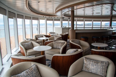 Observation lounge filled with cream upholstered chairs set around small coffee tables in front of wrap around floor to ceiling windows.
