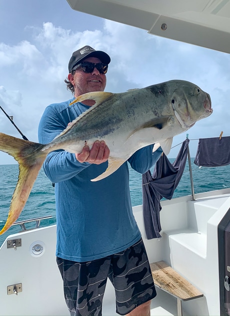 A happy male fisherman stands on a Belize sailboat holding a deep sea fish he caught while on board.