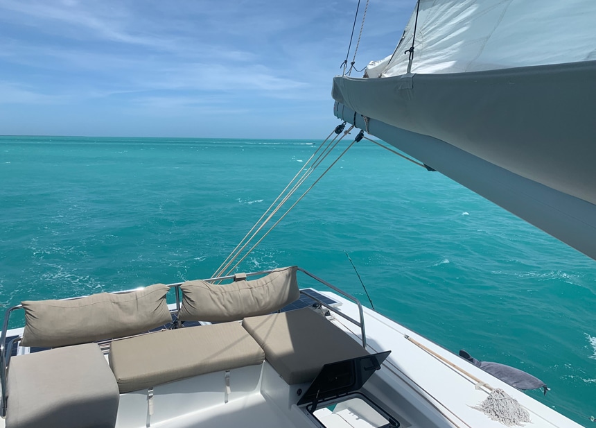 A catamaran cruises the bright teal ocean of Belize. A white boat with tan seat cushions on the top deck and a white sailing mast.