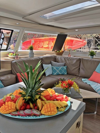 Indoor lounge area aboard Belize catamaran. Upholstered bench seating around a coffee table and a kitchen islands with a plate fresh fruit.