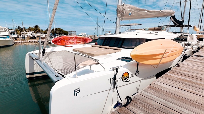 On a blue sky day a bright white catamaran is tied to a wooden dock in Belize, carrying two orange and red kayaks on the deck.