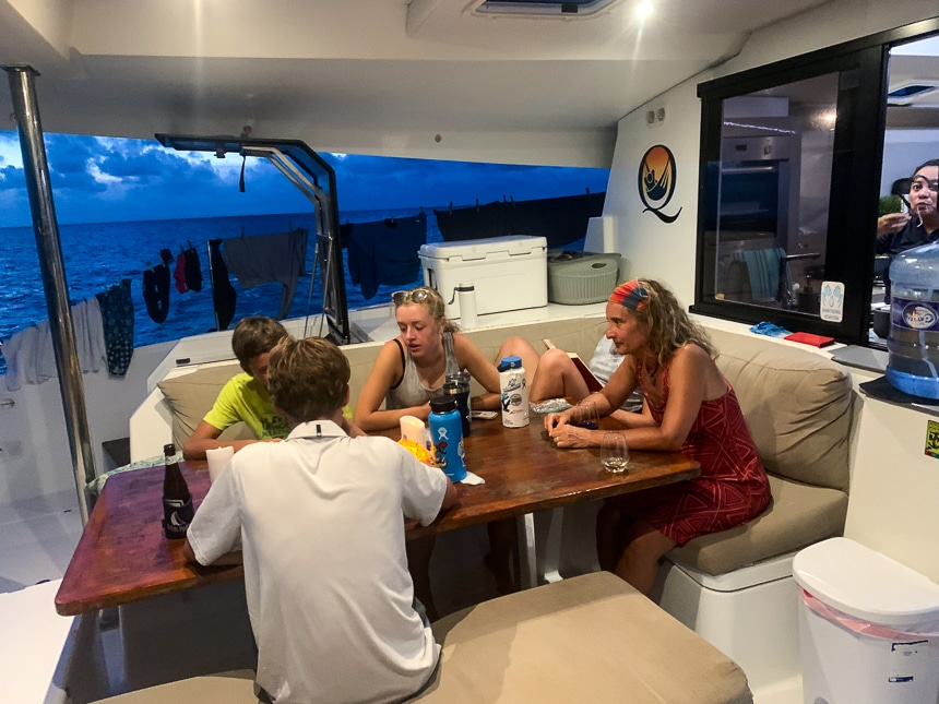 Four people sit around the outside dining table of a Belize sailboat playing games as the sun sets in the distance.