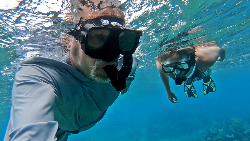An underwater photo of two snorkelers wearing fins, masks and snorkels swim in the shallow blue ocean water of Belize looking for wildlife amongst the coral.