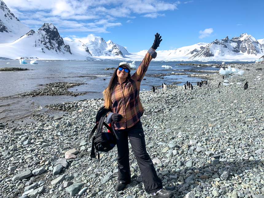 Female AdventureSmith staff poses on grey rocky beach in Antarctica surrounded by penguins and snowy mountain range,