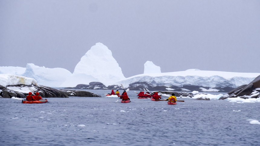 Group of kayakers in red parkas and red double kayaks paddle in the dark waters of Antarctica around icebergs on an overcast day.