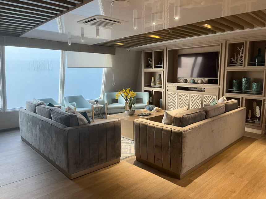 Indoor lounge aboard Endemic Galapagos cruise ship, grey velvet couches with teal upholstered chairs, coffee table and flat screen TV