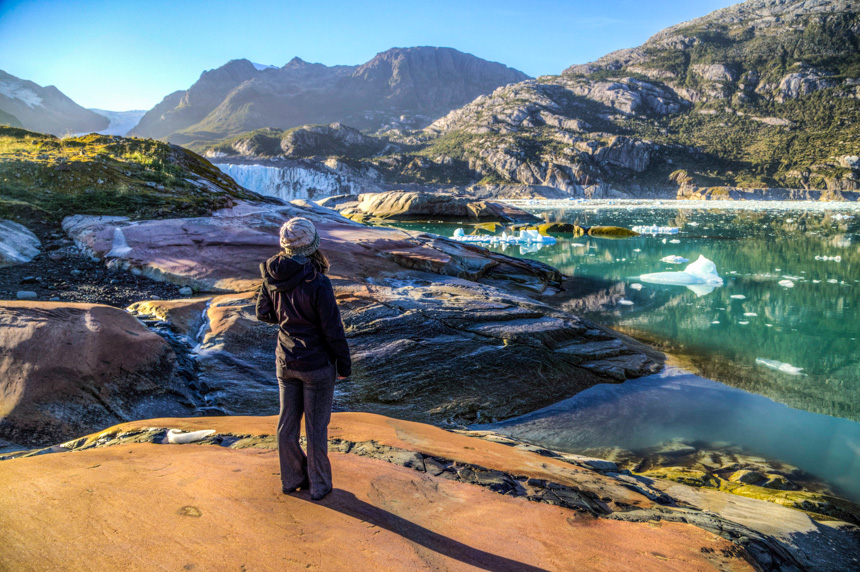 A woman in a black jacket and beanie looks out over a green lake with icebergs and a glacier in Patagonia
