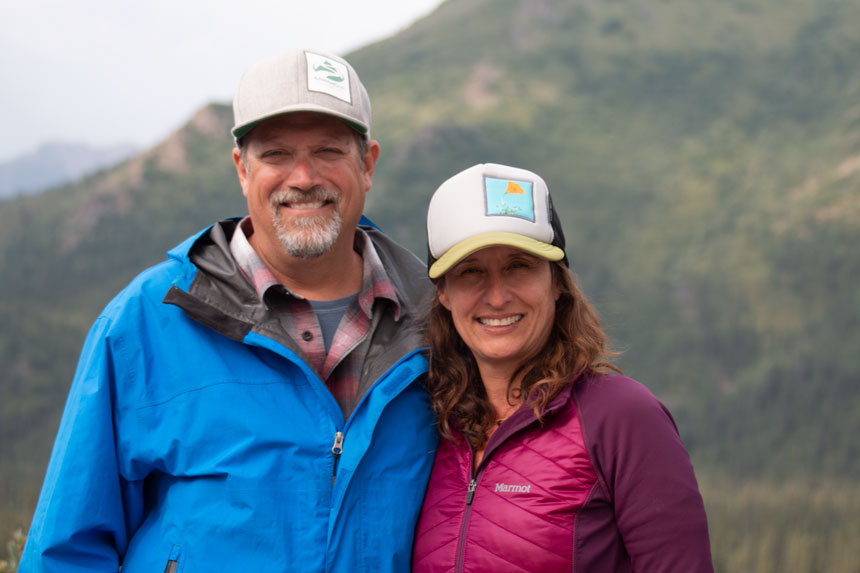 Man in blue jacket u0026 gray hat stands with woman in berry-colored coat u0026 light cap in Denali, Alaska.