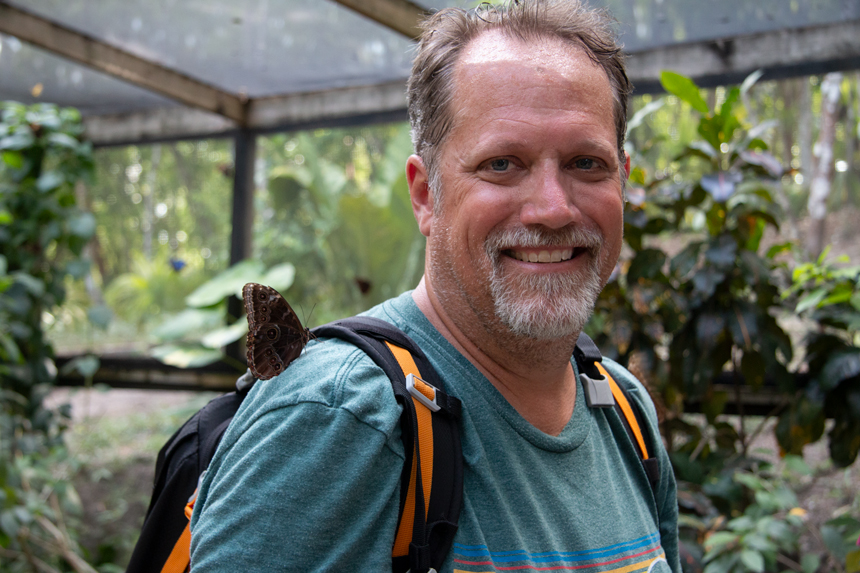 Man in green shirt with backpack smiles as a butterfly rests on his shoulder while traveling in Belize.