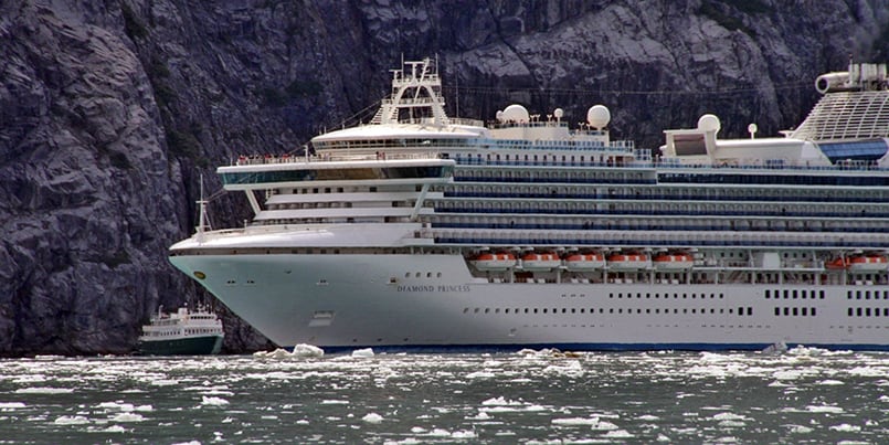A big cruise ship Diamond Princess towers over small ship Wilderness Explorer in front of a rocky cliff with icebergs in the water