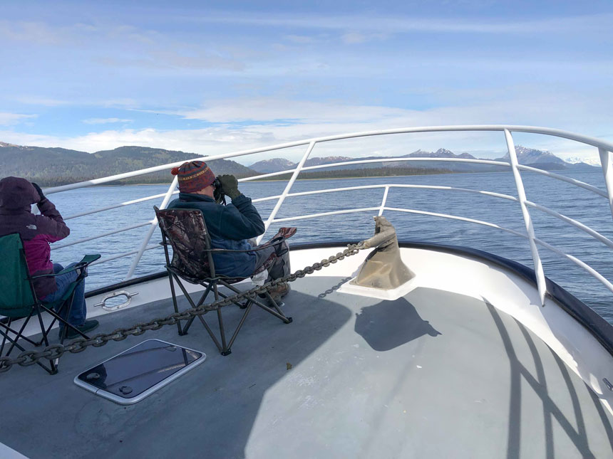 A man sits in a camp chair on the bow of a ship, holding binoculars looking out to the horizon