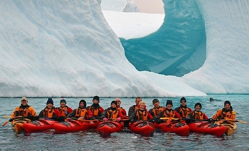 Small ship cruise guests on kayaking excursion lined up together in front of unique shaped and bright glacier.