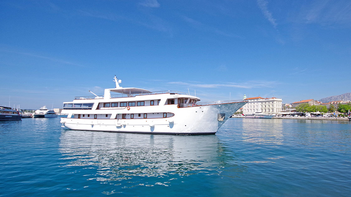 Aurora yacht with 4 guest decks & white exterior sits in calm turquoise water just offshore under sunny skies in Croatia.