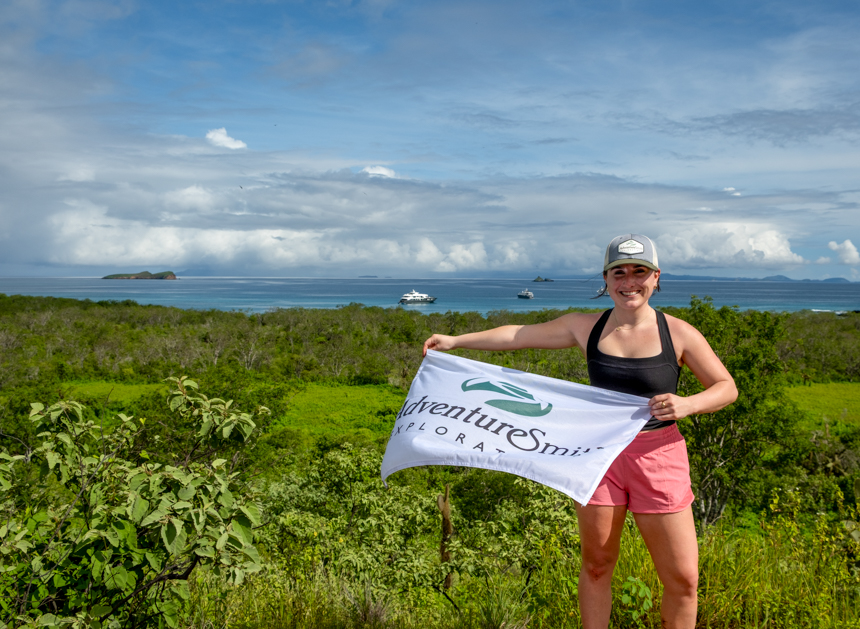 AdventureSmith Grace standing at the top of a lookout spot with an adventuresmith white flag flying with small ships off in the distance
