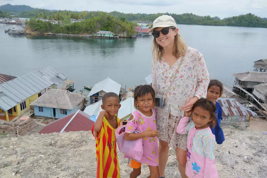 Woman in AdventureSmith baseball cap stands with local children overlooking rustic, colorful seaside homes in Southeast Asia.