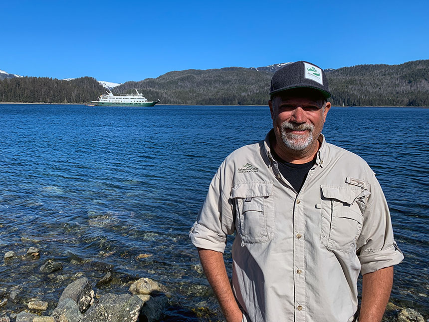 A man in a grey hat and tan button up shirt stands on the shoreline with sunny skies and a small ship behind him