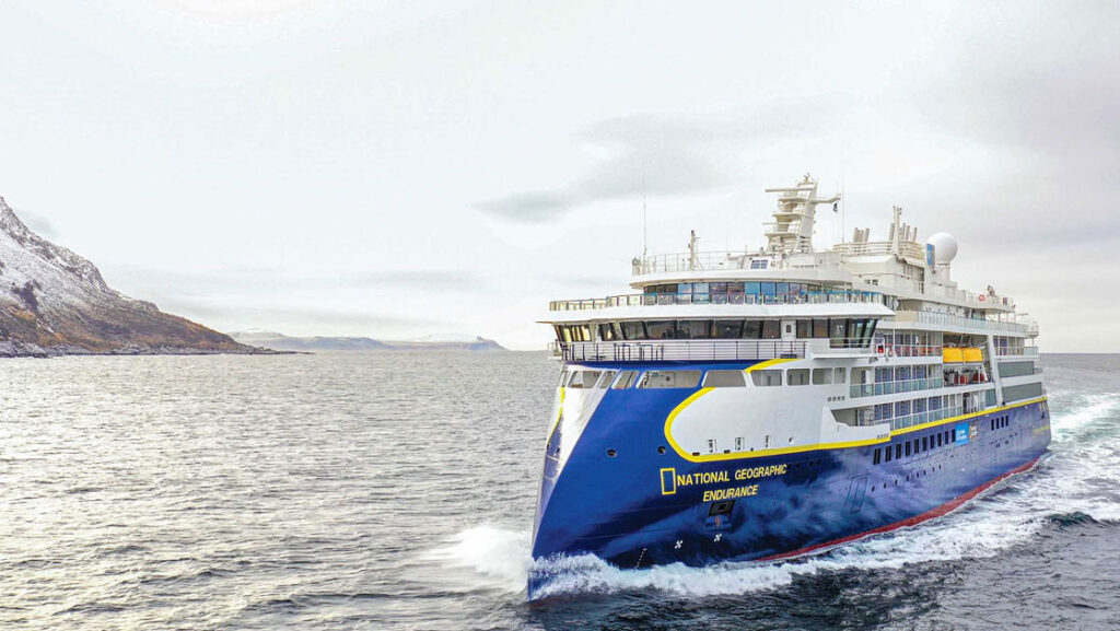 Sleek blue & white National Geographic Endurance expedition ship with multiple decks cruises through pale water on a gray day.